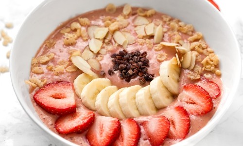 A starwberry and banana smoothie bowl with nuts and cut fruit layered ontop